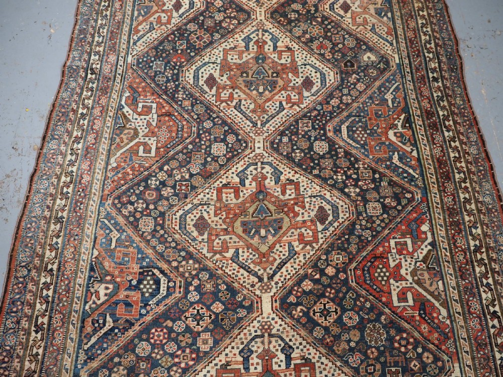 An antique rectangular wool rug featuring three diamond-shaped medallions along a central dark blue field surrounded by geometric borders in terracotta, cream, and blue tones. View IV