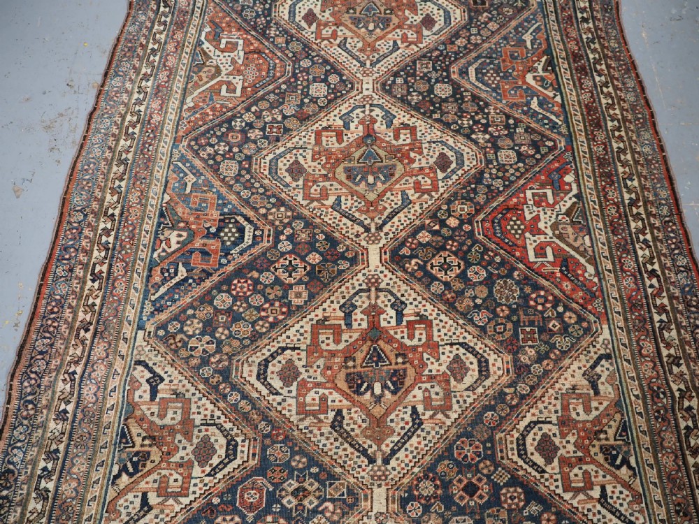 An antique rectangular wool rug featuring three diamond-shaped medallions along a central dark blue field surrounded by geometric borders in terracotta, cream, and blue tones. View V