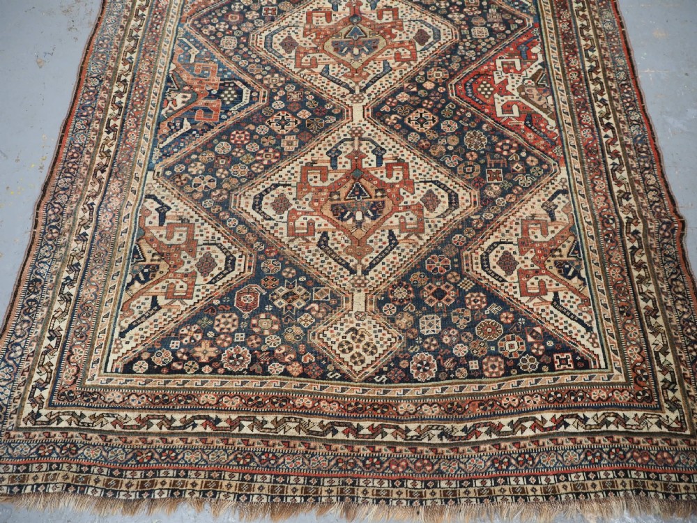An antique rectangular wool rug featuring three diamond-shaped medallions along a central dark blue field surrounded by geometric borders in terracotta, cream, and blue tones. View VI