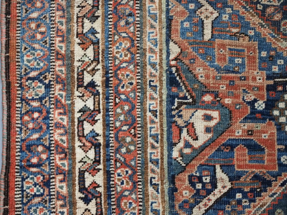 An antique rectangular wool rug featuring three diamond-shaped medallions along a central dark blue field surrounded by geometric borders in terracotta, cream, and blue tones. View VIII