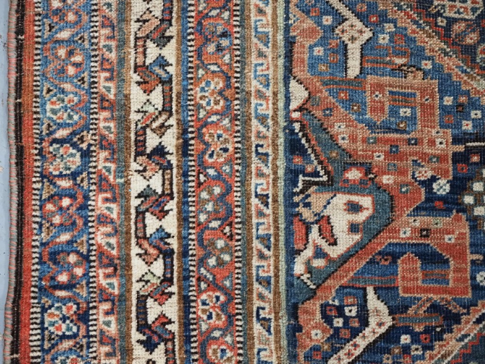 An antique rectangular wool rug featuring three diamond-shaped medallions along a central dark blue field surrounded by geometric borders in terracotta, cream, and blue tones. View IX