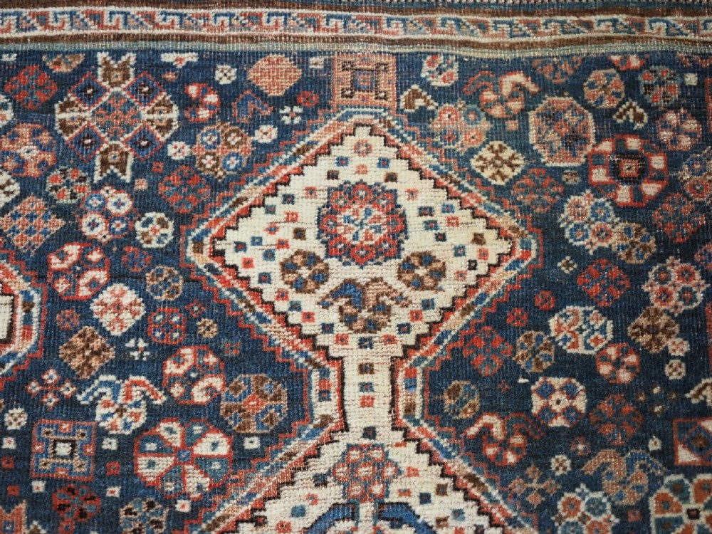 An antique rectangular wool rug featuring three diamond-shaped medallions along a central dark blue field surrounded by geometric borders in terracotta, cream, and blue tones. View X
