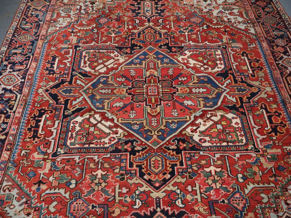 An antique hand-knotted Heriz wool carpet featuring a large geometric central medallion in navy blue, rust red, and cream on a patterned field. View IV