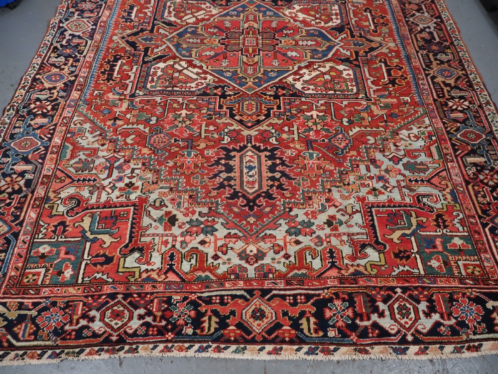 An antique hand-knotted Heriz wool carpet featuring a large geometric central medallion in navy blue, rust red, and cream on a patterned field. View V