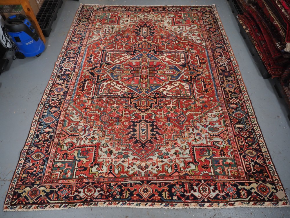 An antique hand-knotted Heriz wool carpet featuring a large geometric central medallion in navy blue, rust red, and cream on a patterned field. View XIV