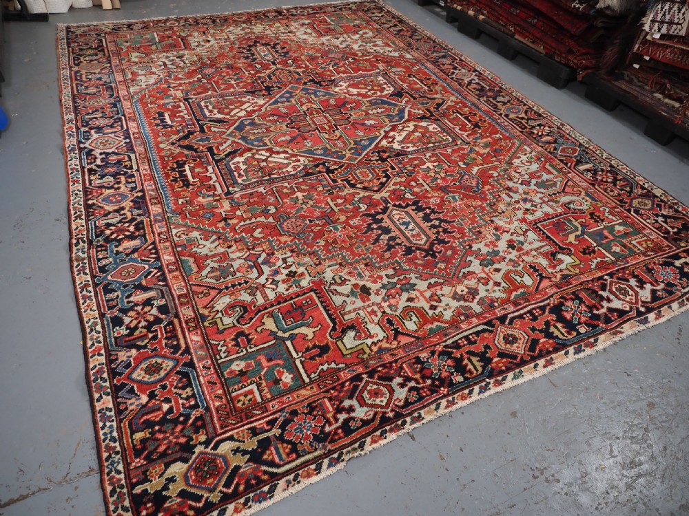 An antique hand-knotted Heriz wool carpet featuring a large geometric central medallion in navy blue, rust red, and cream on a patterned field. View XV