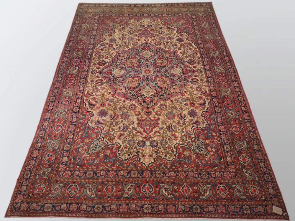 An antique rectangular Isfahan carpet featuring a detailed central medallion, cream field with red and blue floral arabesques, and a decorative multi-banded border.