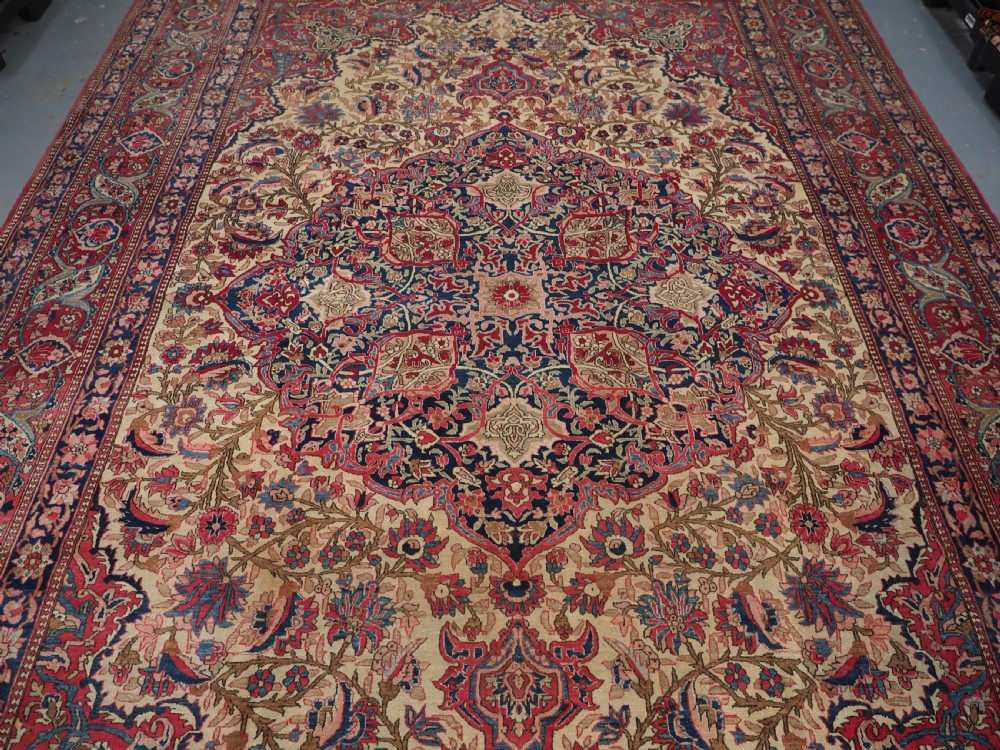 An antique rectangular Isfahan carpet featuring a detailed central medallion, cream field with red and blue floral arabesques, and a decorative multi-banded border. View IV