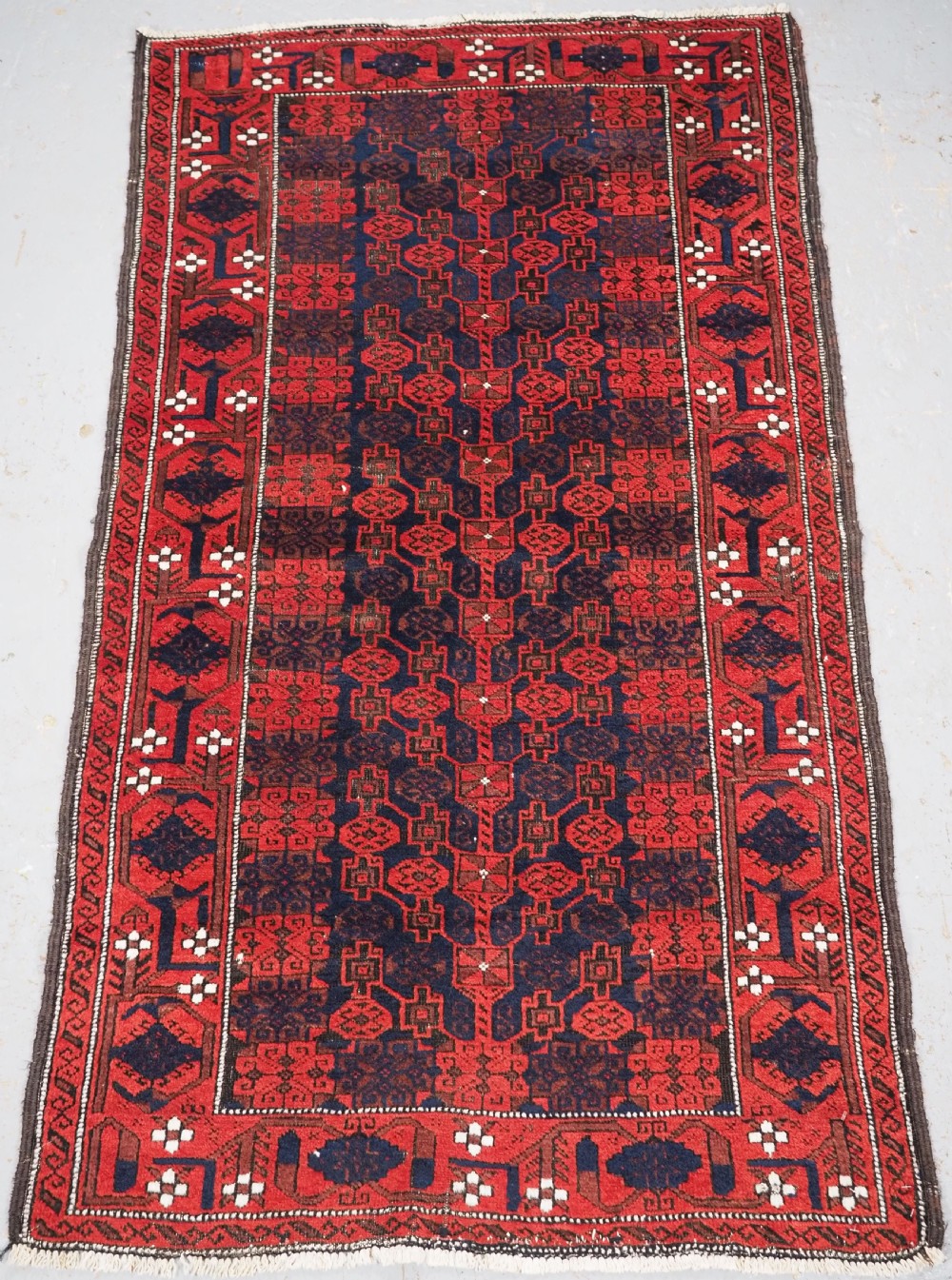 An antique hand-knotted Baluch wool rug featuring a dark blue central field with repeating red geometric motifs inside a patterned red border.