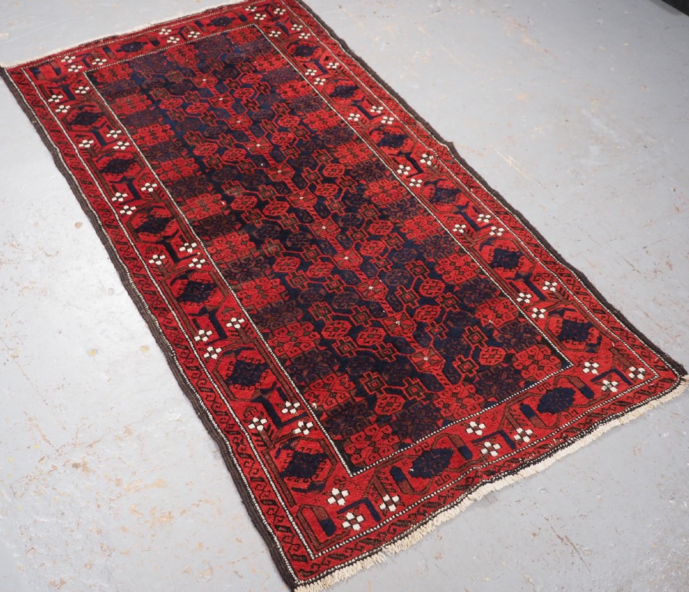 An antique hand-knotted Baluch wool rug featuring a dark blue central field with repeating red geometric motifs inside a patterned red border. View II