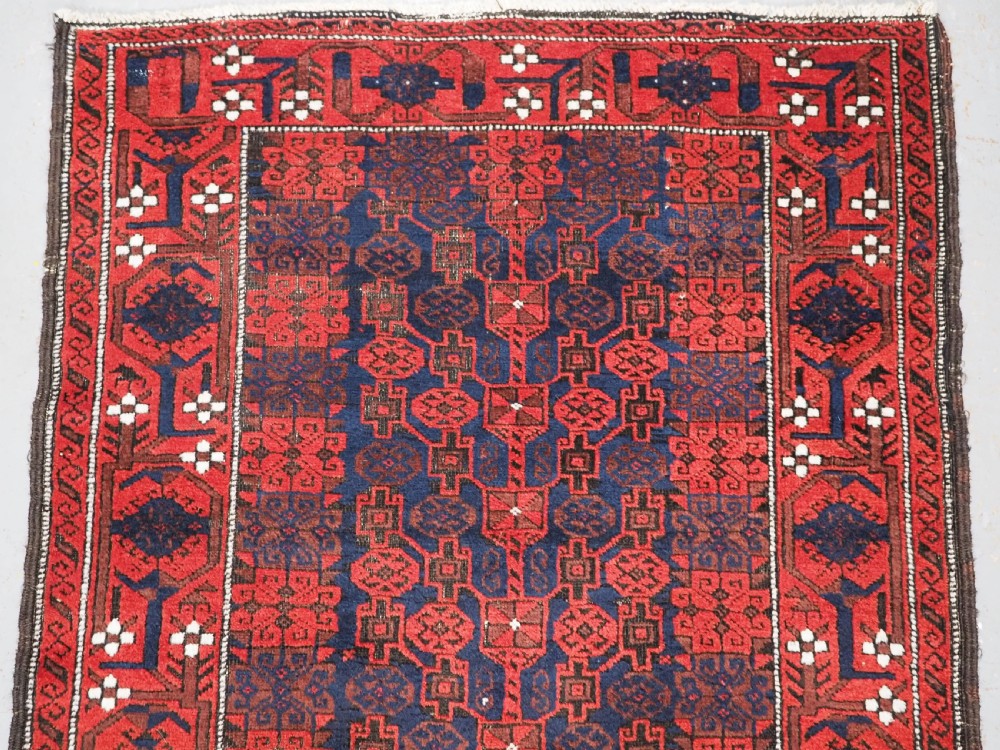 An antique hand-knotted Baluch wool rug featuring a dark blue central field with repeating red geometric motifs inside a patterned red border. View III
