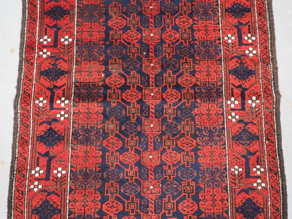 An antique hand-knotted Baluch wool rug featuring a dark blue central field with repeating red geometric motifs inside a patterned red border. View IV