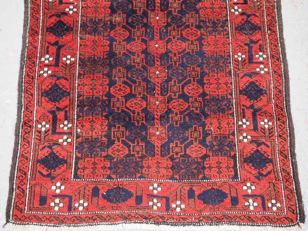 An antique hand-knotted Baluch wool rug featuring a dark blue central field with repeating red geometric motifs inside a patterned red border. View V