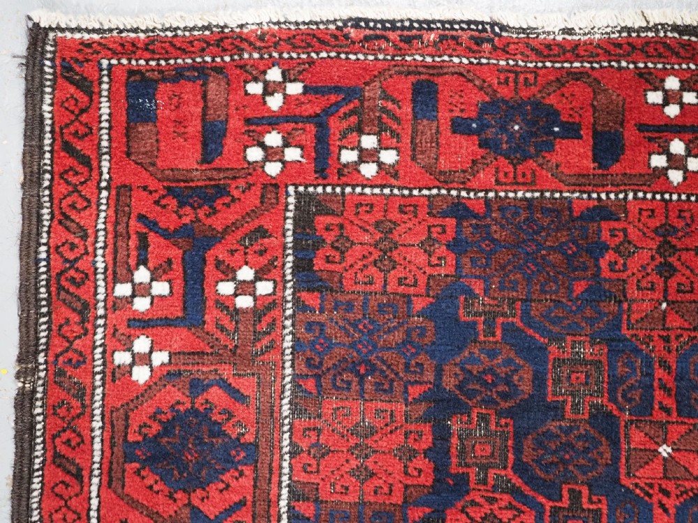 An antique hand-knotted Baluch wool rug featuring a dark blue central field with repeating red geometric motifs inside a patterned red border. View VI