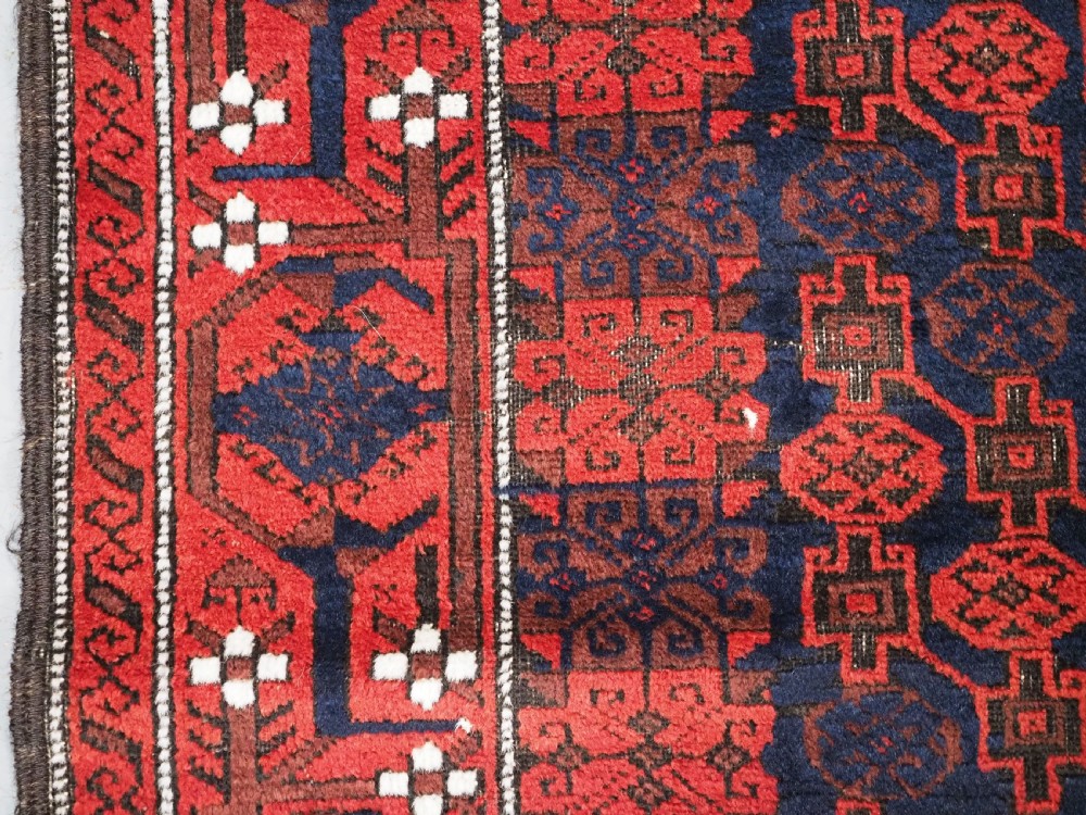 An antique hand-knotted Baluch wool rug featuring a dark blue central field with repeating red geometric motifs inside a patterned red border. View VII