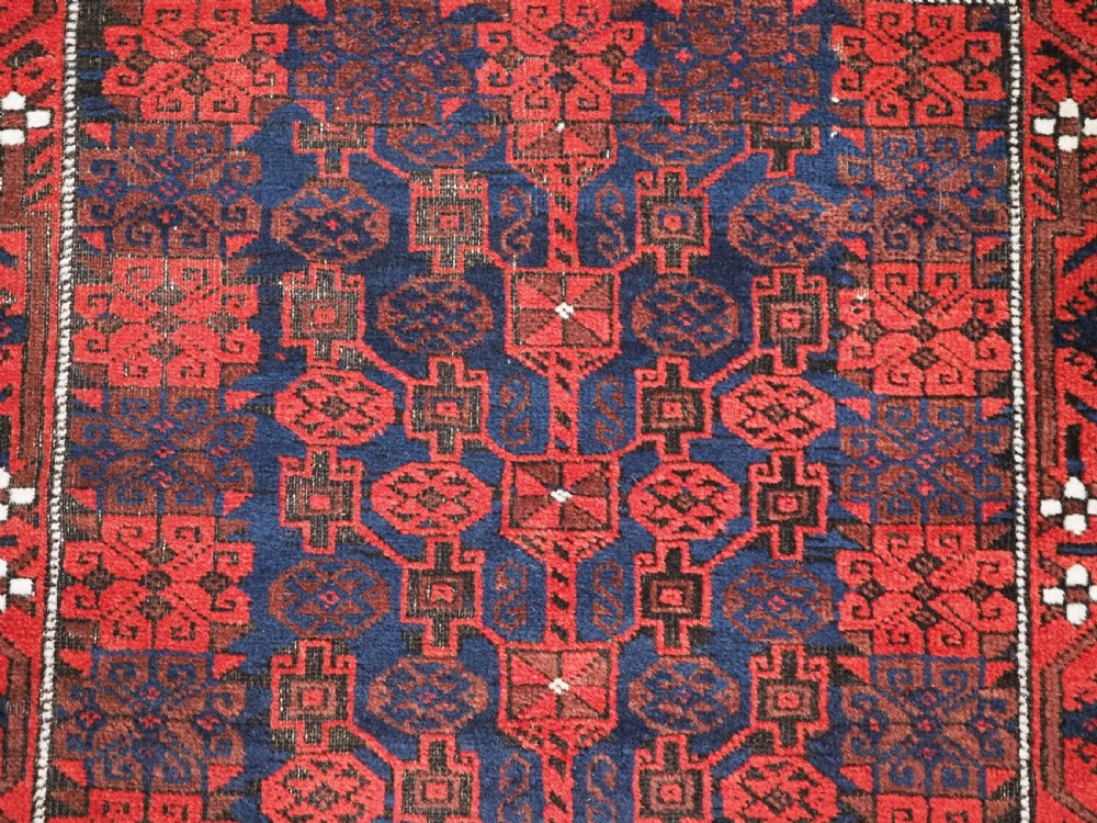 An antique hand-knotted Baluch wool rug featuring a dark blue central field with repeating red geometric motifs inside a patterned red border. View VIII