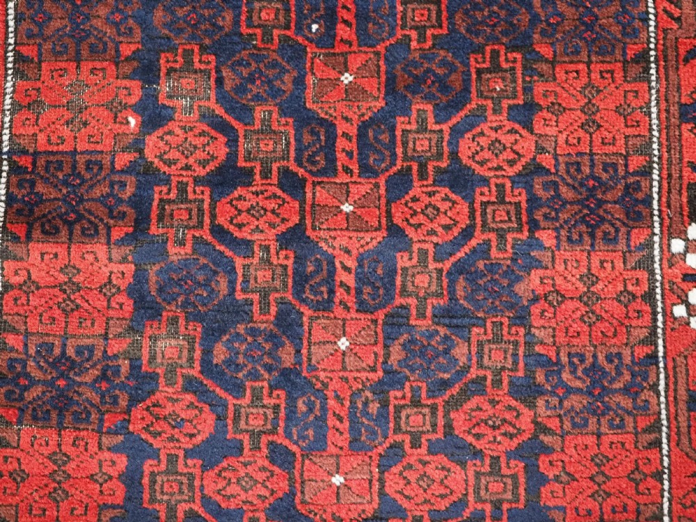 An antique hand-knotted Baluch wool rug featuring a dark blue central field with repeating red geometric motifs inside a patterned red border. View IX