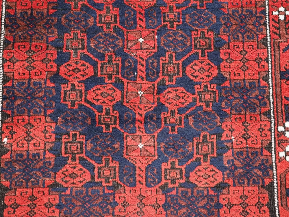 An antique hand-knotted Baluch wool rug featuring a dark blue central field with repeating red geometric motifs inside a patterned red border. View X