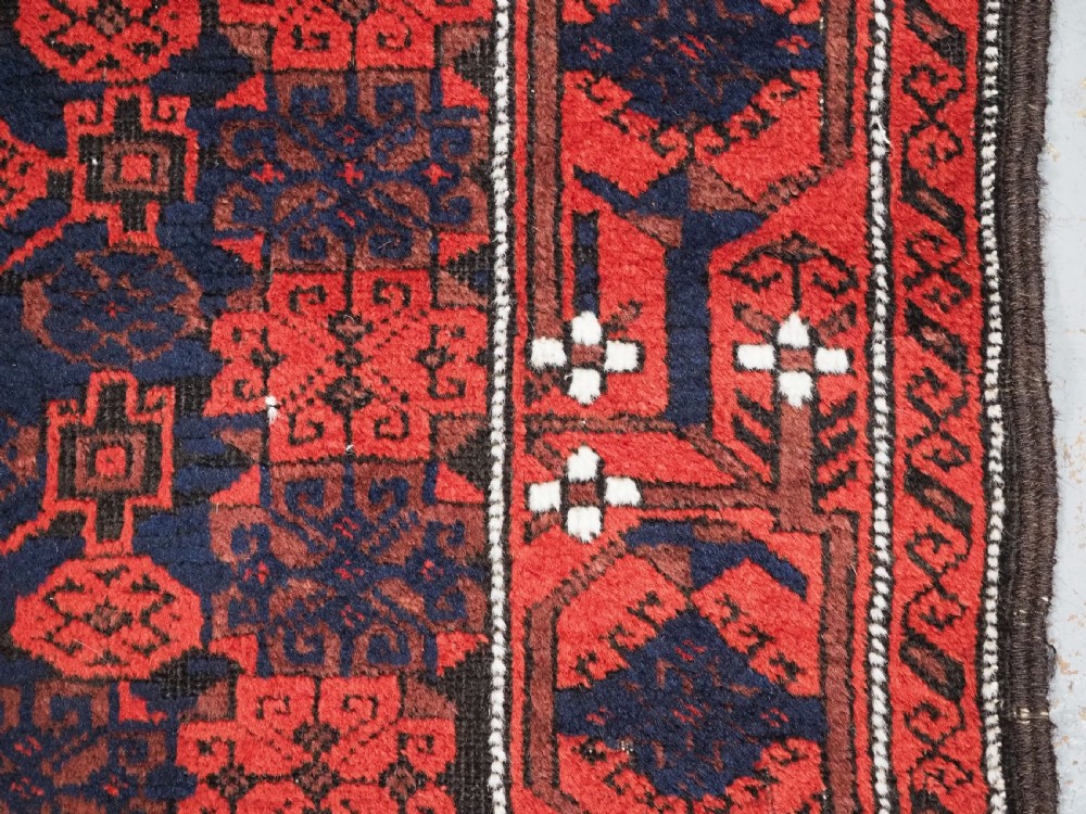 An antique hand-knotted Baluch wool rug featuring a dark blue central field with repeating red geometric motifs inside a patterned red border. View XI