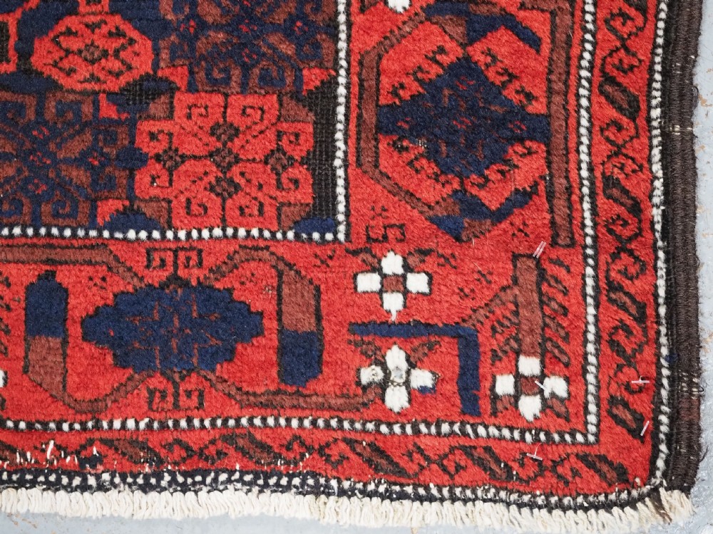 An antique hand-knotted Baluch wool rug featuring a dark blue central field with repeating red geometric motifs inside a patterned red border. View XII