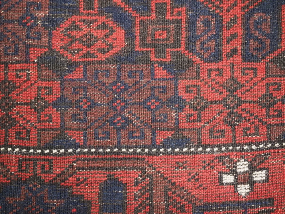 An antique hand-knotted Baluch wool rug featuring a dark blue central field with repeating red geometric motifs inside a patterned red border. View XIII