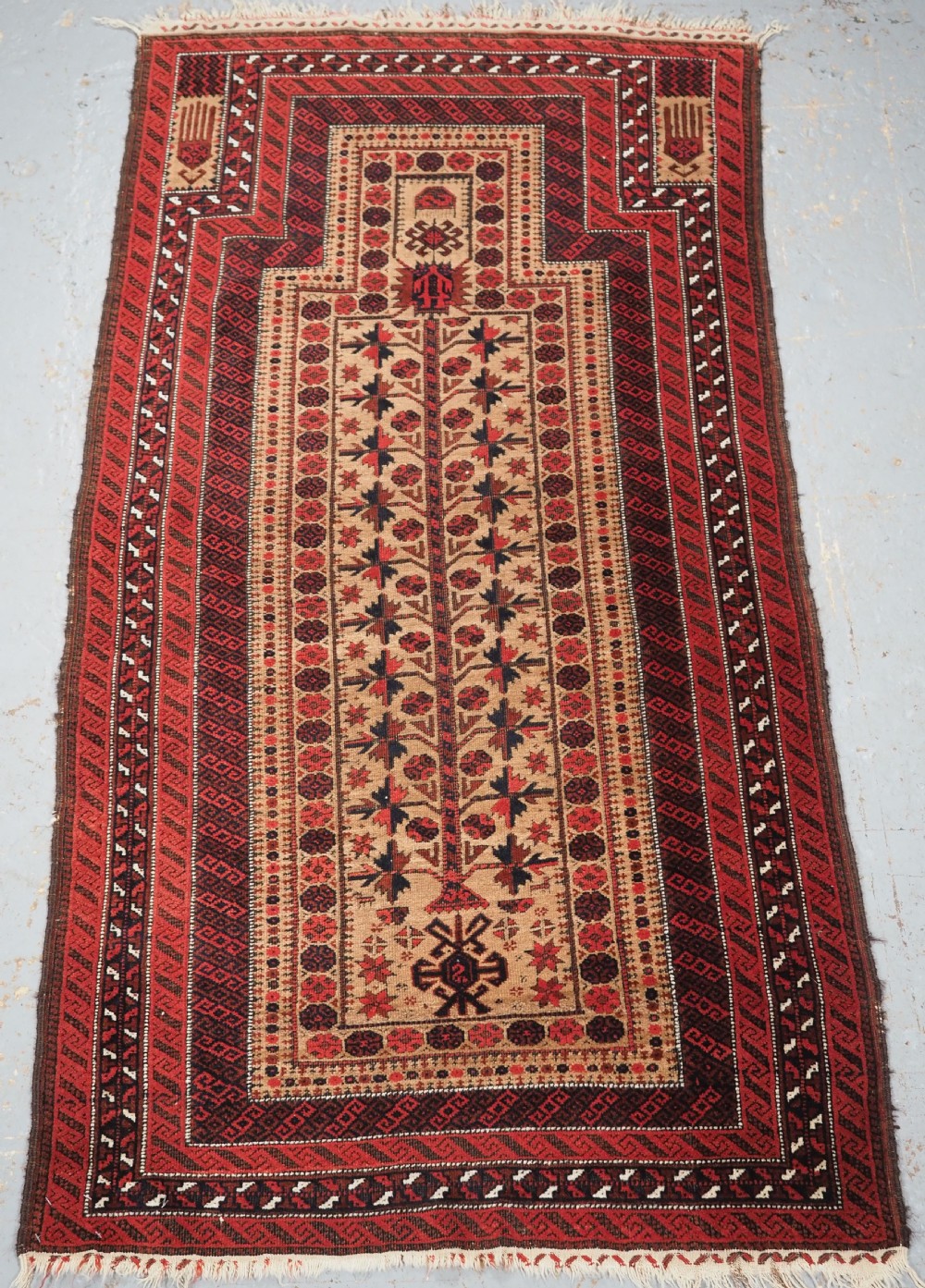 An antique Baluch wool prayer rug featuring a central mihrab arch, tree motifs, hand icons, and geometric borders in dark red, brown, and camel shades.