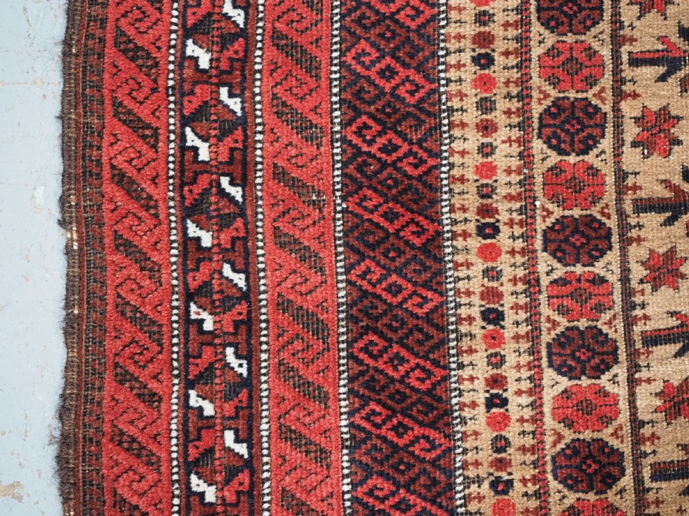 An antique Baluch wool prayer rug featuring a central mihrab arch, tree motifs, hand icons, and geometric borders in dark red, brown, and camel shades. View VII