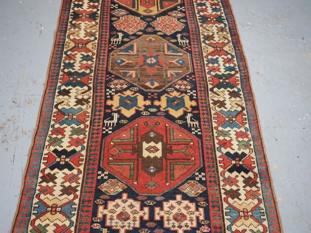 A long antique hand-knotted Caucasian wool runner rug featuring repeating geometric medallions in red, navy blue, ochre, and cream. View III