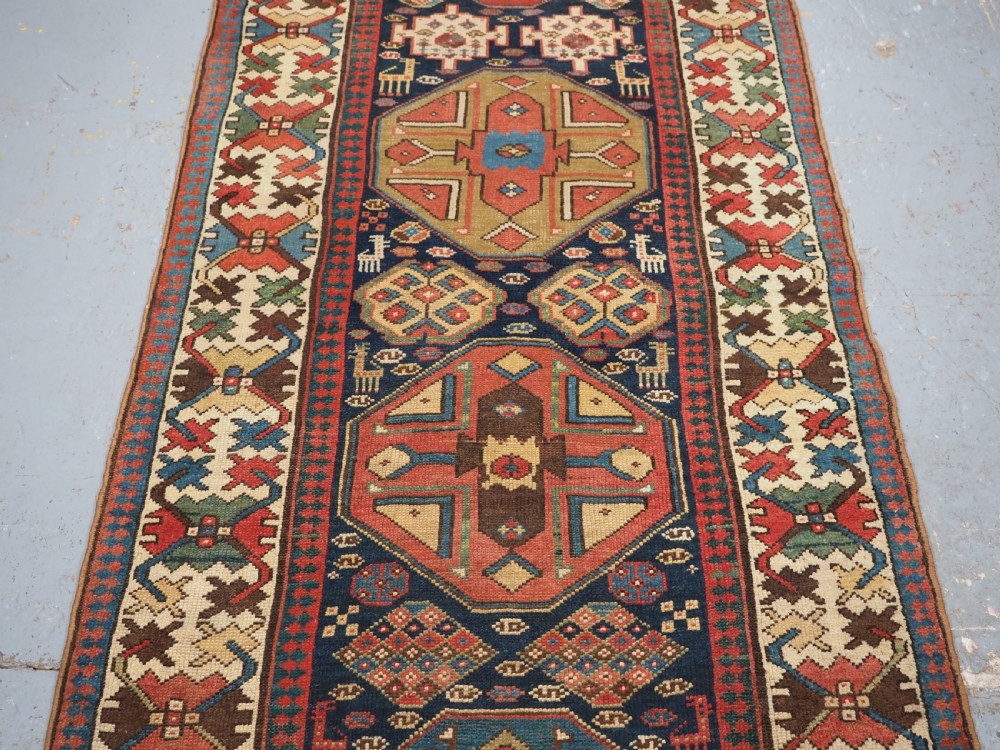 A long antique hand-knotted Caucasian wool runner rug featuring repeating geometric medallions in red, navy blue, ochre, and cream. View IV