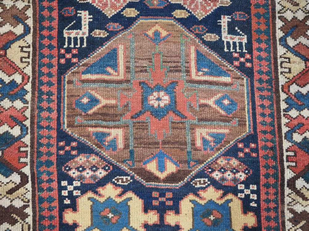 A long antique hand-knotted Caucasian wool runner rug featuring repeating geometric medallions in red, navy blue, ochre, and cream. View IX