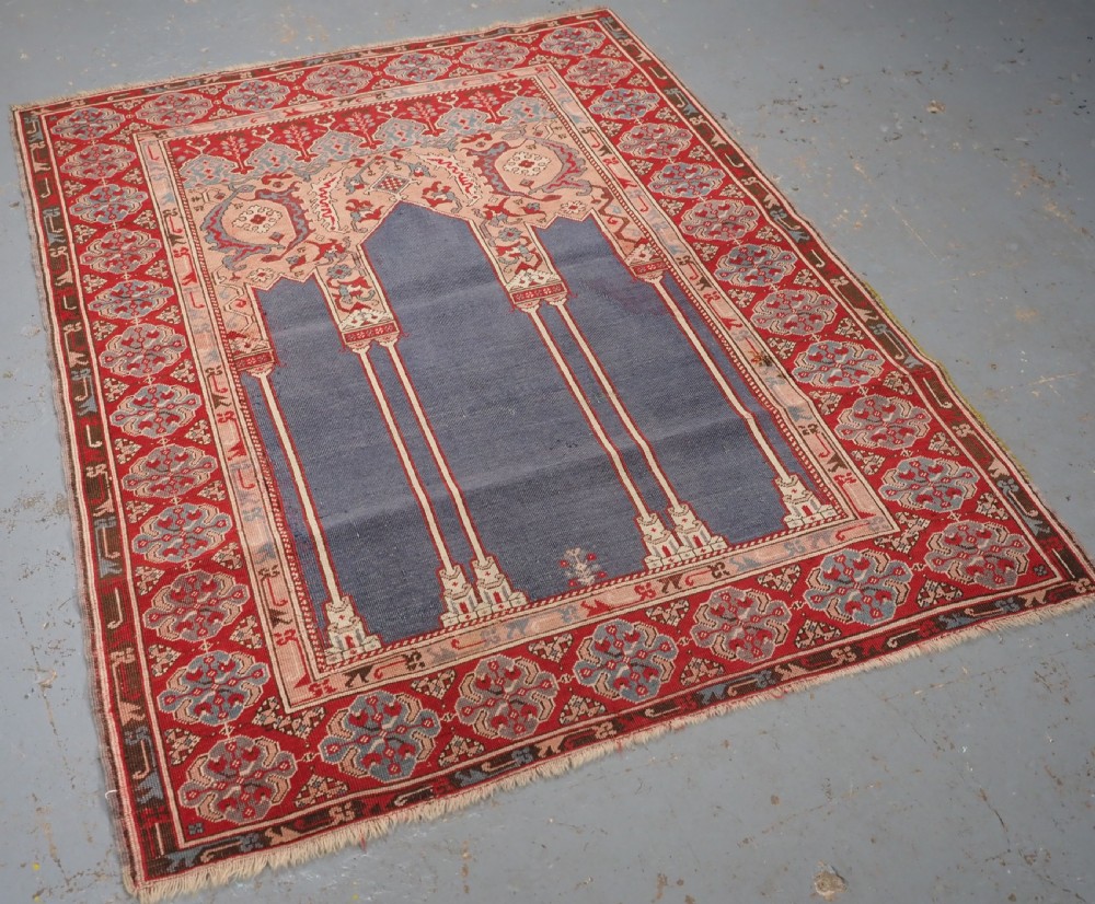 A hand-knotted wool prayer rug with a blue central column niche motif framed by beige spandrels and a red geometric border. View II