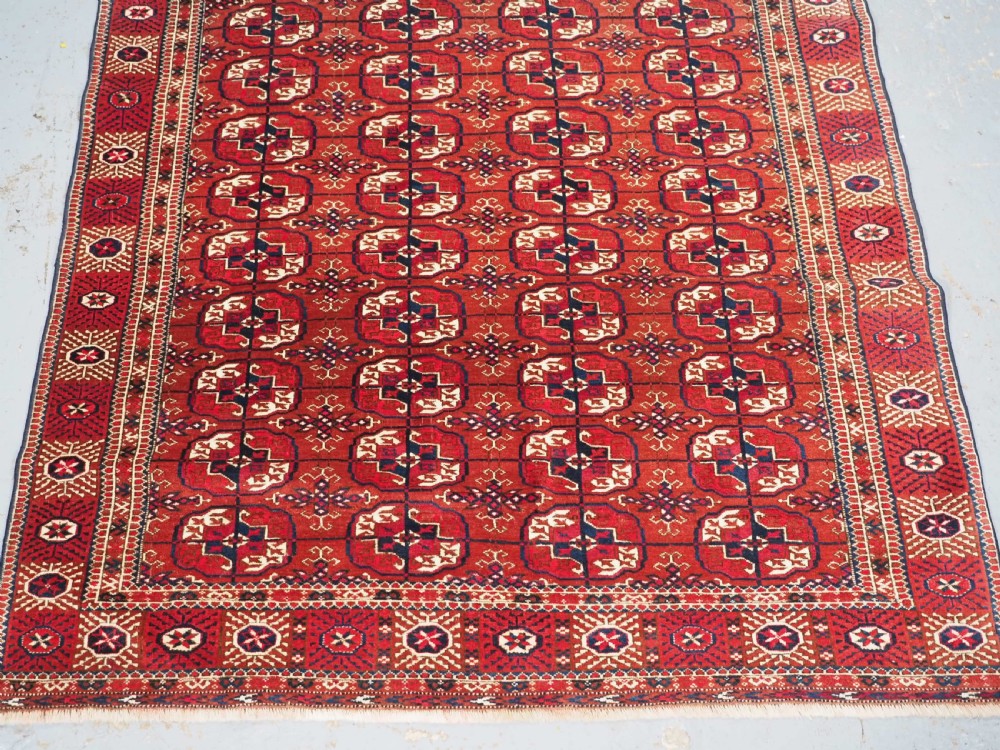 A rectangular hand-knotted Tekke Turkmen wool rug featuring repeated geometric primary guls on a deep red field surrounded by multi-band borders. View IV