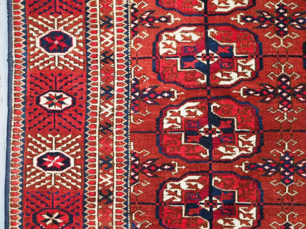 A rectangular hand-knotted Tekke Turkmen wool rug featuring repeated geometric primary guls on a deep red field surrounded by multi-band borders. View VI