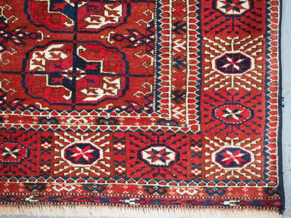 A rectangular hand-knotted Tekke Turkmen wool rug featuring repeated geometric primary guls on a deep red field surrounded by multi-band borders. View X
