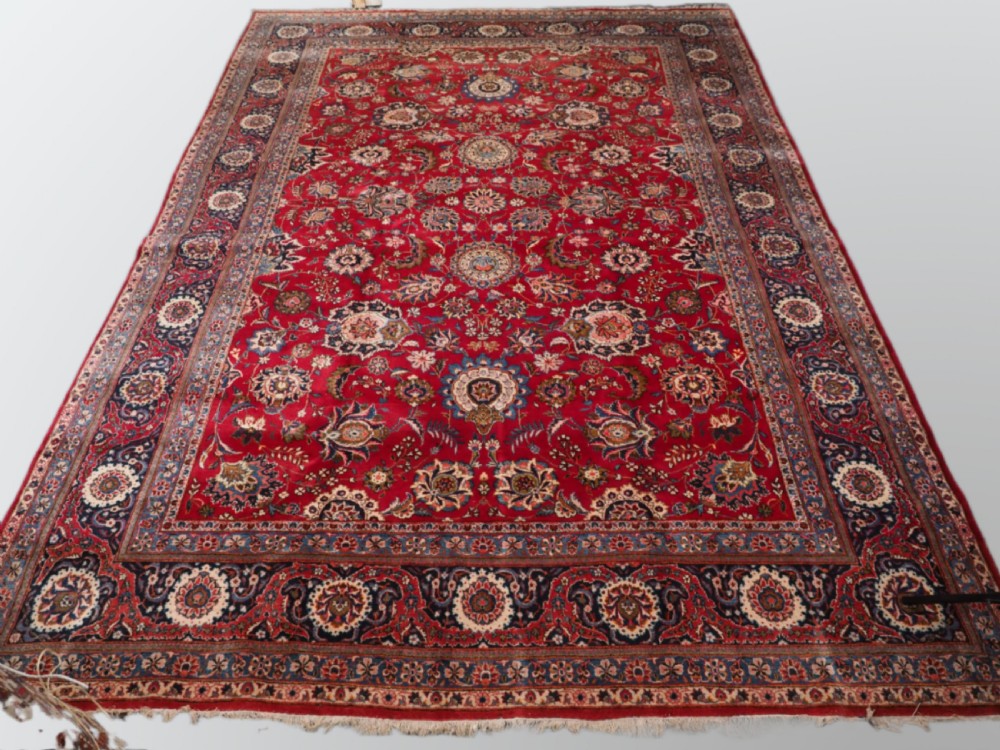 A large hand-knotted Kashan wool carpet featuring an intricate all-over floral pattern on a rich red field framed by a navy blue patterned border.