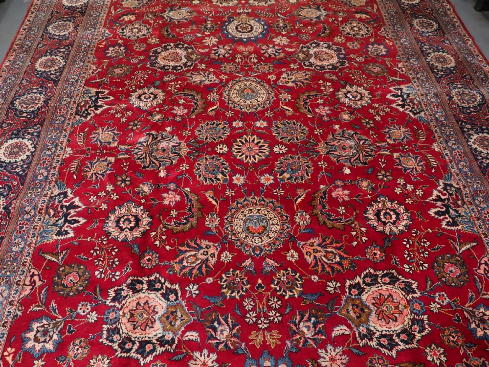 A large hand-knotted Kashan wool carpet featuring an intricate all-over floral pattern on a rich red field framed by a navy blue patterned border. View V