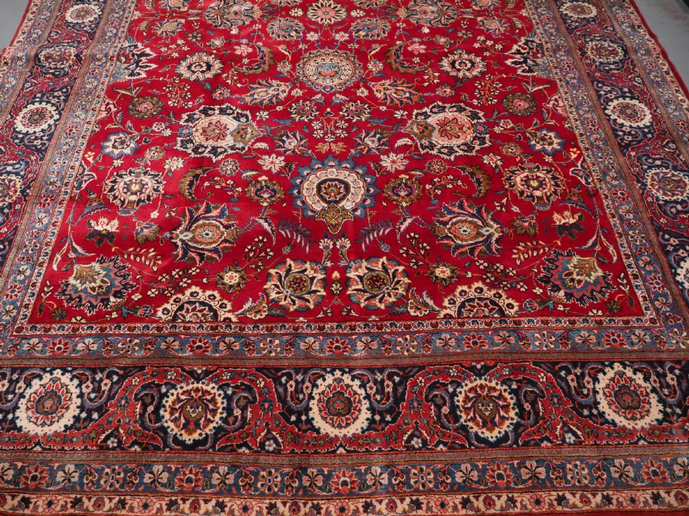 A large hand-knotted Kashan wool carpet featuring an intricate all-over floral pattern on a rich red field framed by a navy blue patterned border. View VI