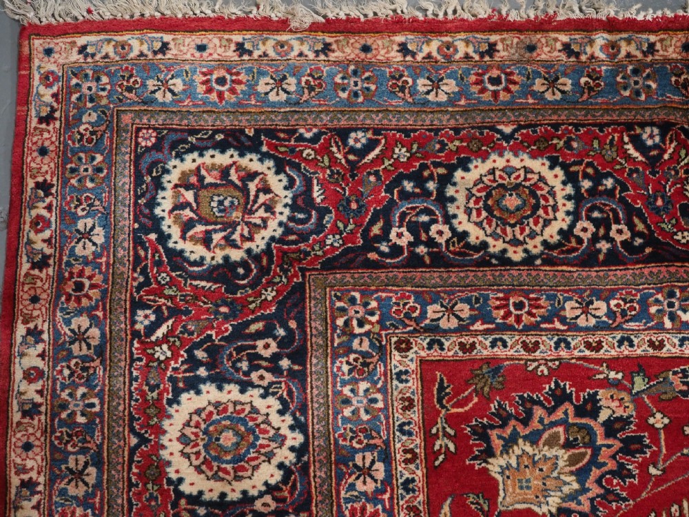 A large hand-knotted Kashan wool carpet featuring an intricate all-over floral pattern on a rich red field framed by a navy blue patterned border. View VII