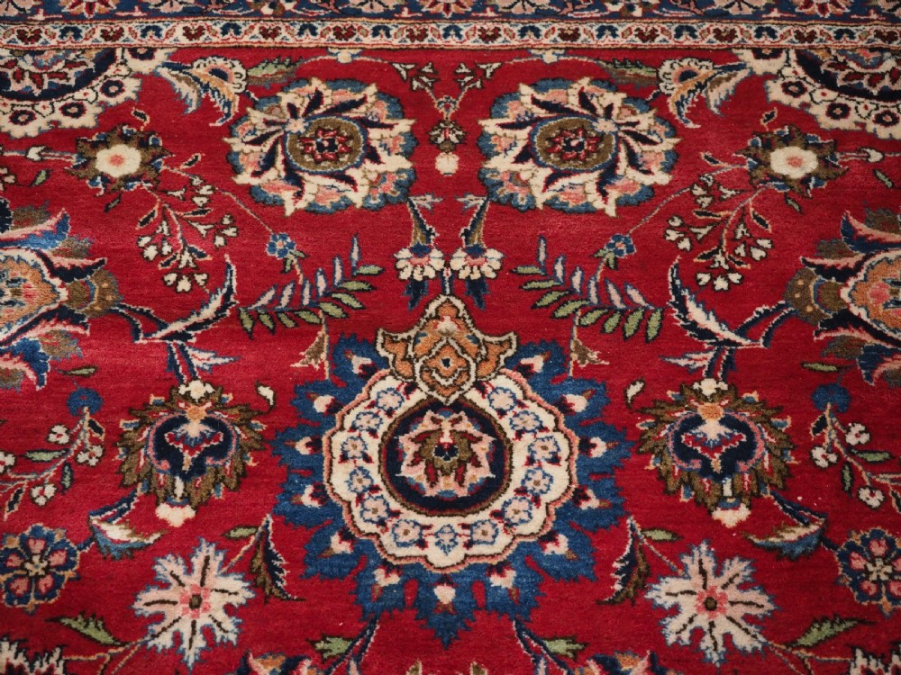 A large hand-knotted Kashan wool carpet featuring an intricate all-over floral pattern on a rich red field framed by a navy blue patterned border. View IX
