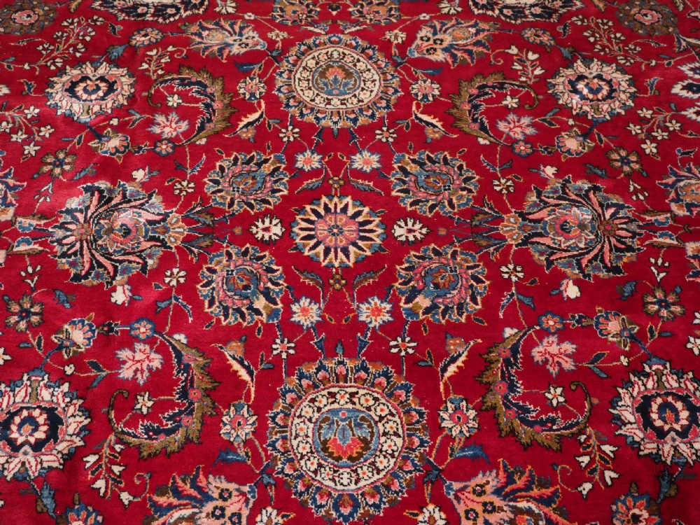 A large hand-knotted Kashan wool carpet featuring an intricate all-over floral pattern on a rich red field framed by a navy blue patterned border. View X