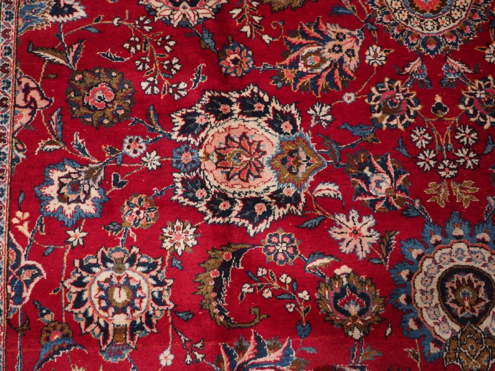 A large hand-knotted Kashan wool carpet featuring an intricate all-over floral pattern on a rich red field framed by a navy blue patterned border. View XII
