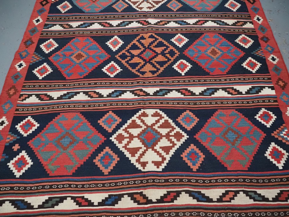 An antique Shahsavan wool flatweave kilim rug featuring geometric diamond motifs in red, navy blue, terracotta, and light blue tones. View V