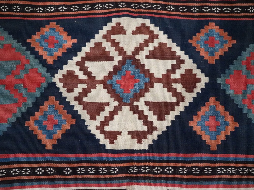 An antique Shahsavan wool flatweave kilim rug featuring geometric diamond motifs in red, navy blue, terracotta, and light blue tones. View X