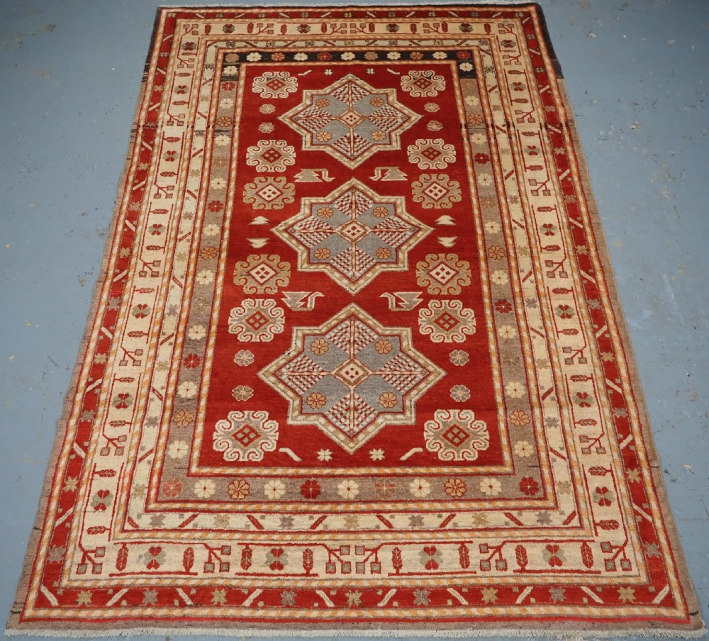 An antique East Turkestan Khotan wool area rug featuring three light blue geometric star medallions on a red field within wide cream and beige patterned borders.