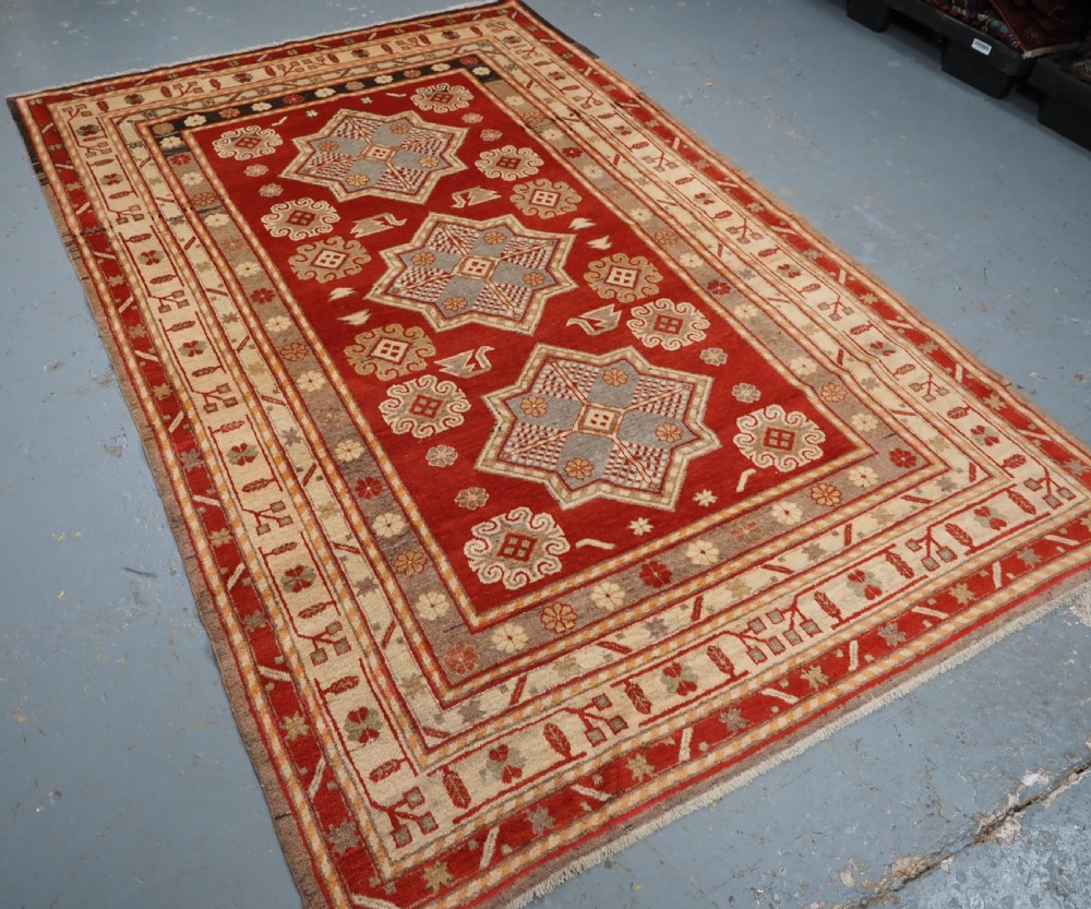 An antique East Turkestan Khotan wool area rug featuring three light blue geometric star medallions on a red field within wide cream and beige patterned borders. View II