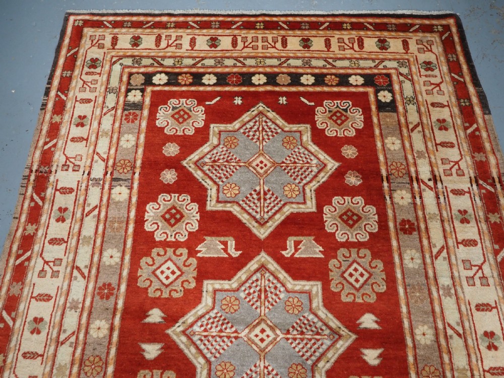 An antique East Turkestan Khotan wool area rug featuring three light blue geometric star medallions on a red field within wide cream and beige patterned borders. View III