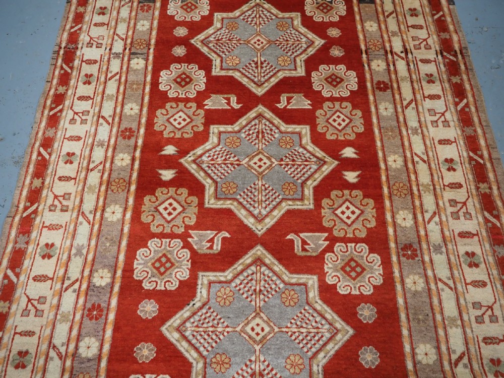 An antique East Turkestan Khotan wool area rug featuring three light blue geometric star medallions on a red field within wide cream and beige patterned borders. View IV