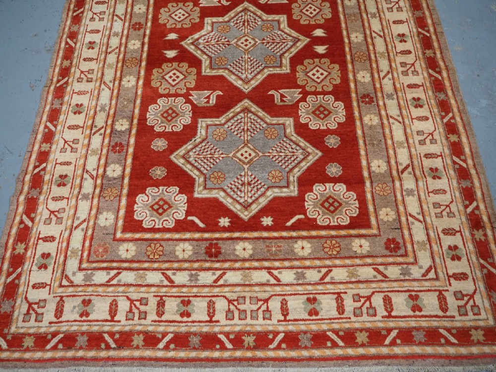 An antique East Turkestan Khotan wool area rug featuring three light blue geometric star medallions on a red field within wide cream and beige patterned borders. View V