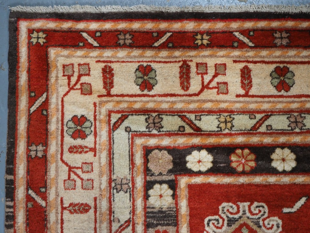 An antique East Turkestan Khotan wool area rug featuring three light blue geometric star medallions on a red field within wide cream and beige patterned borders. View VI