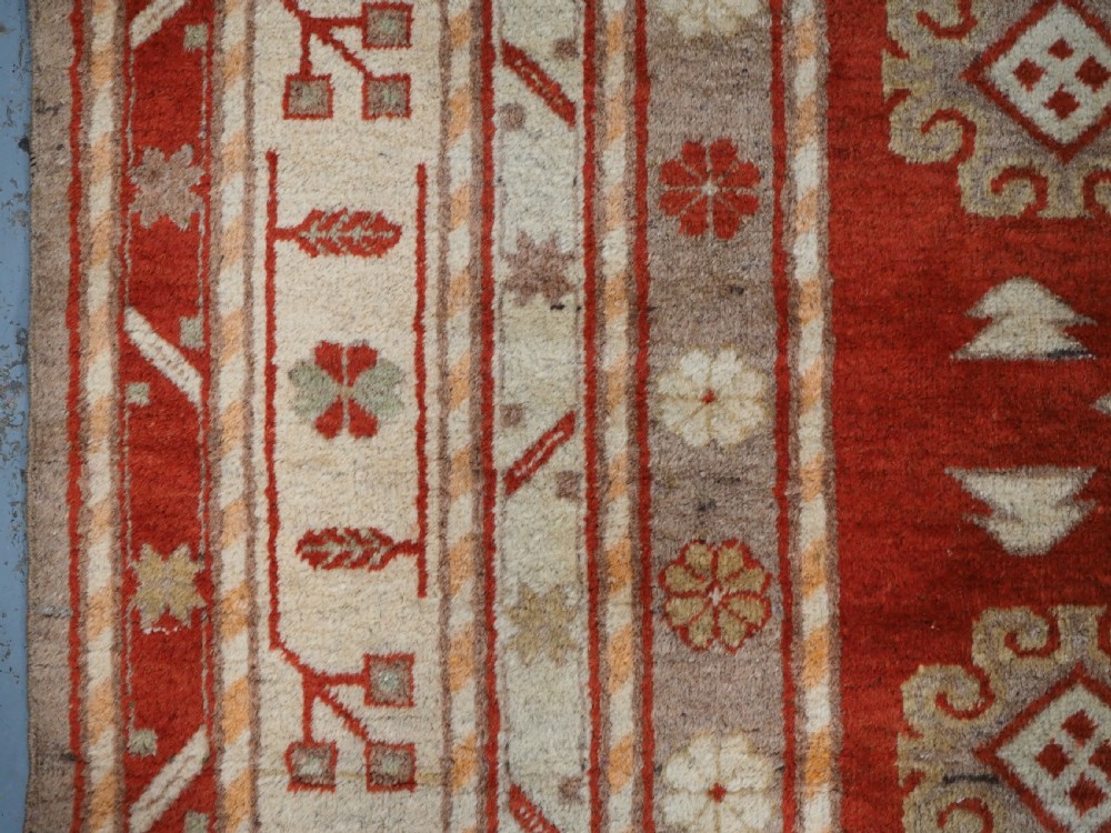 An antique East Turkestan Khotan wool area rug featuring three light blue geometric star medallions on a red field within wide cream and beige patterned borders. View VII